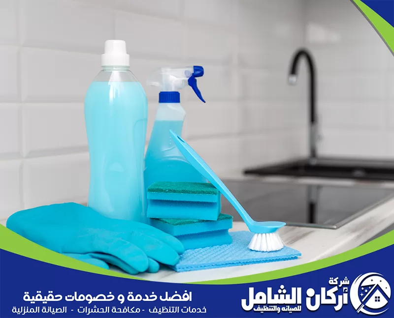أدوات تنظيف مطبخ موضوعة على سطح رخامي، تشمل قفازات، إسفنج، فرشاة، عبوة منظف، وبخاخ تنظيف بجوار حوض المطبخ.