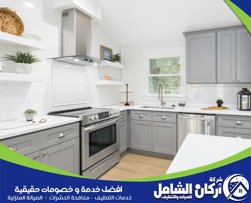 مطبخ حديث ونظيف بتصميم أبيض ورمادي، يحتوي على خزائن مرتبة، فرن، شفاط، حوض، ونافذة جانبية مع لمسات ديكور بسيطة.