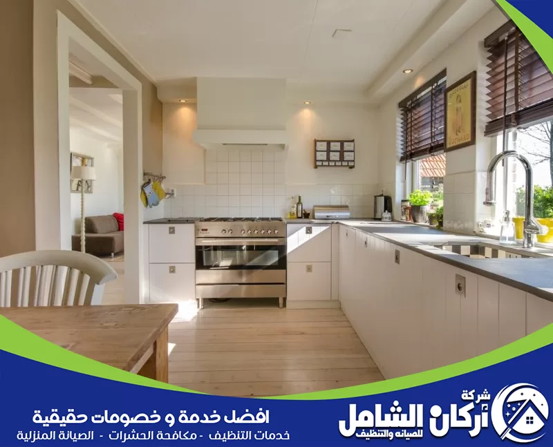 مطبخ واسع ومرتب بتصميم هادئ، يحتوي على خزائن بيضاء، حوض، فرن كبير، نوافذ جانبية، وإضاءة طبيعية واضحة.